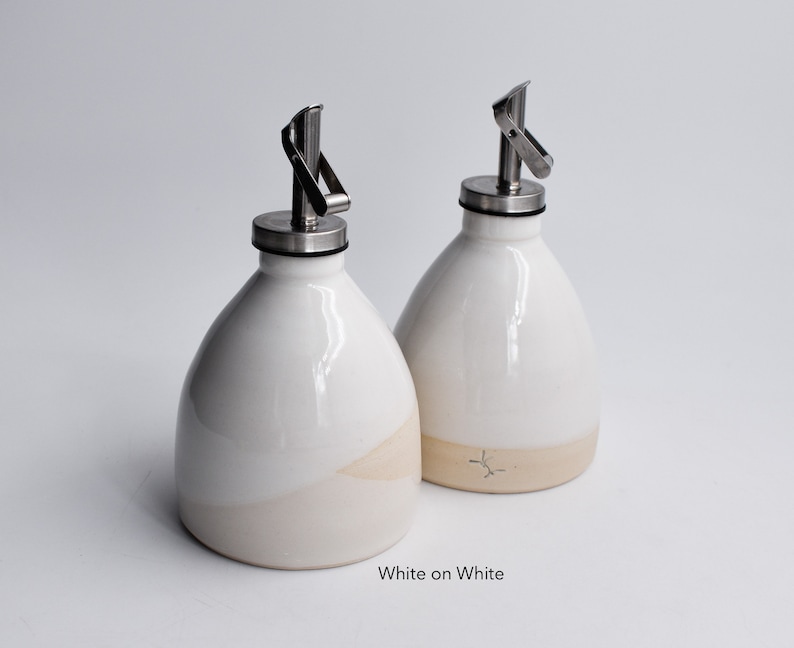 May include: Two white ceramic oil and vinegar bottles with stainless steel pour spouts. The bottles have a white glaze on the top half and a cream glaze on the bottom half. The text "White on White" is printed below the bottles.