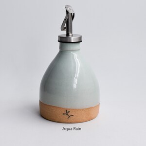 May include: A light blue ceramic oil and vinegar bottle with a stainless steel pour spout and a cork bottom. The bottle has a brown band around the bottom and the text "Aqua Rain" is printed below the bottle.