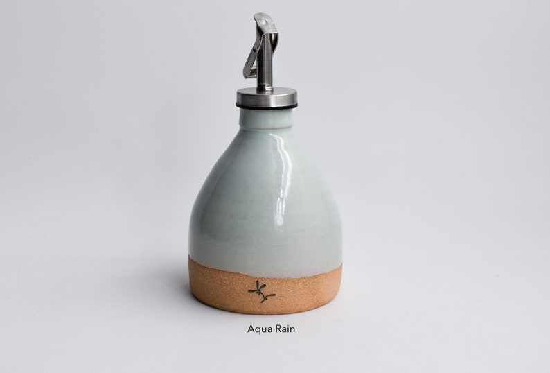 May include: A light blue ceramic oil and vinegar bottle with a stainless steel pour spout and a cork bottom. The bottle has a brown band around the bottom and the text "Aqua Rain" is printed below the bottle.