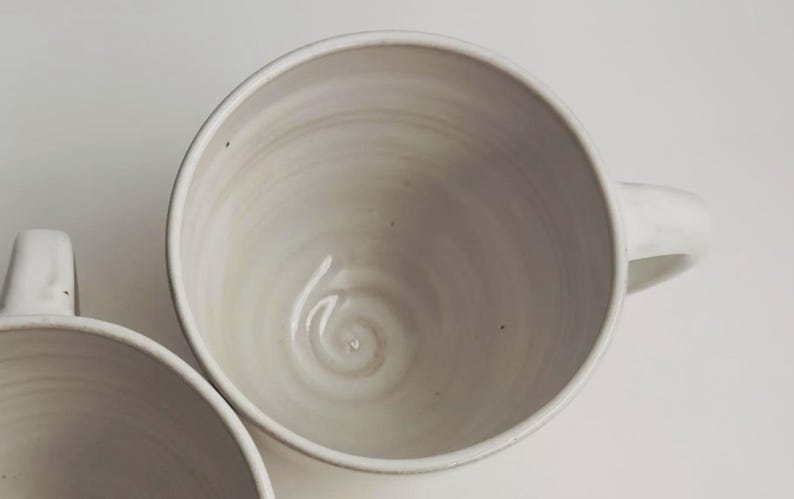 K&ouml;nnte beinhalten: Zwei cremewei&szlig;e Keramikbecher mit glatter, gl&auml;nzender Oberfl&auml;che. Das Innere jedes Bechers weist ein dezentes, wirbelndes Muster auf, das auf der T&ouml;pferscheibe entstanden ist, mit einem abgerundeten Henkel an der Seite f&uuml;r ein schlichtes Design.