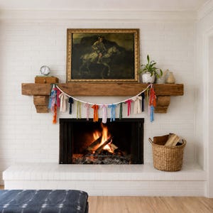 May include: A fireplace with a white brick surround and a wooden mantel. A garland of colourful fabric strips and white beads hangs above the fireplace. A painting of a person on a horse hangs above the mantel.