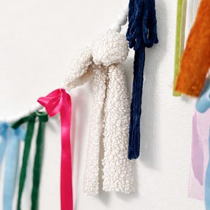 May include: A decorative garland featuring a white, textured, plush bunny-shaped element, connected to a string of wooden beads. Accented with a vibrant pink ribbon and a dark blue velvet ribbon. Other colourful ribbons are also visible.