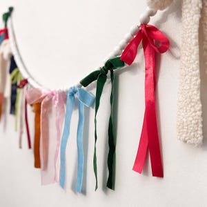 May include: A decorative garland featuring a white circular frame, embellished with a variety of ribbons and wooden beads. The ribbons are in shades of pink, blue, green, and red, creating a bright and cheerful decoration. The garland is set against a white wall.