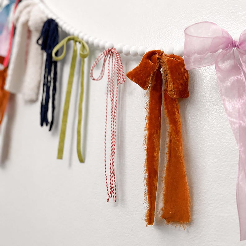 May include: A decorative garland with white wooden beads and colourful ribbons and bows. The ribbons are in shades of orange, green, navy, and pink. The garland is suitable for festive occasions.