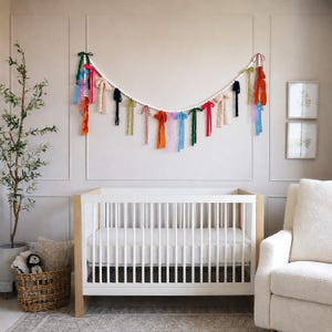 May include: A white cot with a light wood frame is the focal point. Above, a garland of white beads and colourful ribbons hangs on the wall. A wicker basket with a stuffed toy and a potted plant are in the corner.
