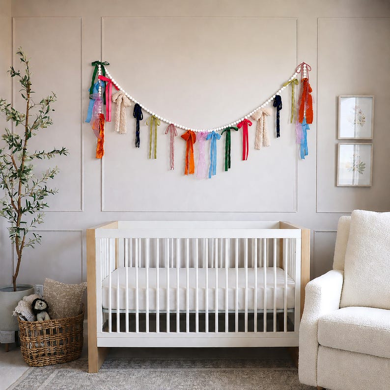 May include: A white cot with a light wood frame is the focal point. Above, a garland of white beads and colourful ribbons hangs on the wall. A wicker basket with a stuffed toy and a potted plant are in the corner.