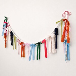 May include: A decorative garland featuring a white beaded string adorned with colourful fabric ribbons. The ribbons are in various shades, including red, blue, green, and orange. The garland is suspended against a white wall.