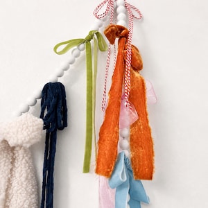May include: A decorative garland with white wooden beads and fabric tassels in various colours. The tassels include navy blue, green, orange, pink, and light blue fabrics. Red and white twine is also used.