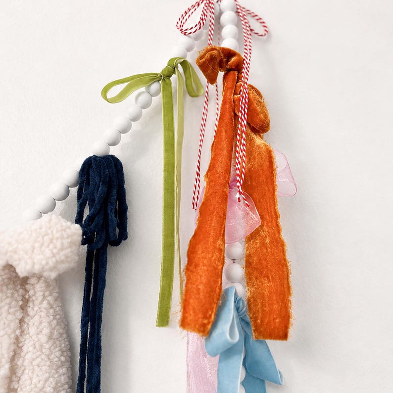 May include: A decorative garland with white wooden beads and fabric tassels in various colours. The tassels include navy blue, green, orange, pink, and light blue fabrics. Red and white twine is also used.