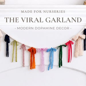 May include: A garland featuring white beads and a variety of colourful ribbons and fabric tassels. The ribbons are in shades of pink, orange, blue, green, and cream. The image text reads "MADE FOR NURSERIES" and "THE VIRAL GARLAND - MODERN DOPAMINE DECOR."