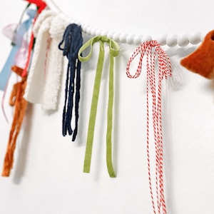 May include: A decorative garland with a selection of colourful ribbons, fabric scraps, and twine. The garland includes navy blue, green, and red accents, strung on a white beaded strand. The background is a plain white wall.