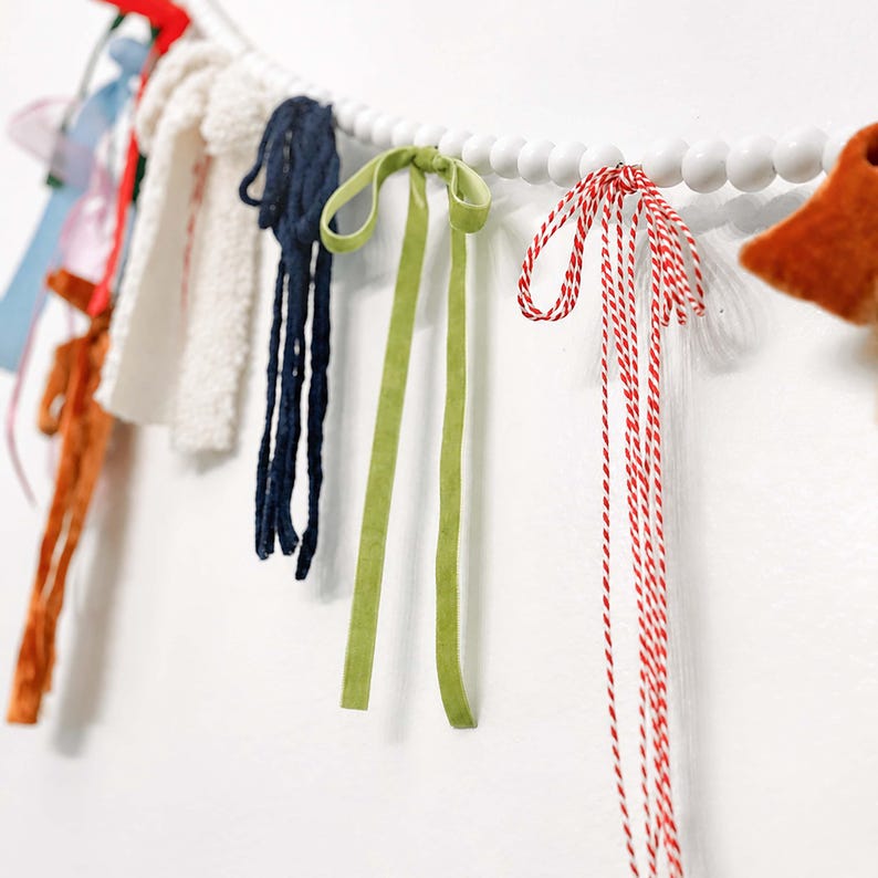 May include: A decorative garland with a selection of colourful ribbons, fabric scraps, and twine. The garland includes navy blue, green, and red accents, strung on a white beaded strand. The background is a plain white wall.
