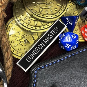 May include: A collection of gold-colored coins with a design of a dragon, a black leather pouch with a blue stitching detail, a silver clamp, a brown rope, and various dice including a red 20-sided die, a blue 20-sided die, a purple 20-sided die, a silver metal die, and a red 6-sided die. A black label with the text "DUNGEON MASTER" is visible.