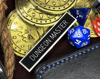 May include: A collection of gold-colored coins with a design of a dragon, a black leather pouch with a blue stitching detail, a silver clamp, a brown rope, and various dice including a red 20-sided die, a blue 20-sided die, a purple 20-sided die, a silver metal die, and a red 6-sided die. A black label with the text "DUNGEON MASTER" is visible.