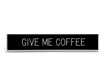 May include: A rectangular black sign with the white text "GIVE ME COFFEE". The sign is set against a white background.