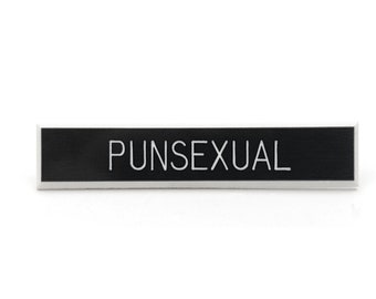 May include: A rectangular black name tag with the word "PUNSEXUAL" in white, centered on the tag. The tag has a thin, silver border and is set against a white background.
