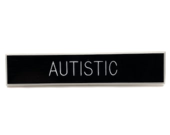 May include: A rectangular black sign with the word "AUTISTIC" in white, centered. The sign has a thin, silver border and is set against a plain white background. The text is in a sans-serif font.