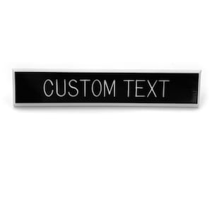 Peut inclure: Une étiquette de nom rectangulaire noire avec une bordure argentée et le texte "CUSTOM TEXT" en blanc.