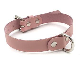 May include: A light pink leather collar with a silver-colored metal buckle and a D-ring. The collar is secured with two silver-colored rivets near the buckle and one near the D-ring. The collar is designed to be adjustable.