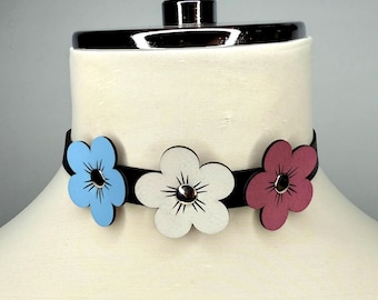 May include: A black choker necklace featuring three flower pendants in blue, white, and pink. Each flower has a silver-colored center. The choker is displayed on a mannequin.