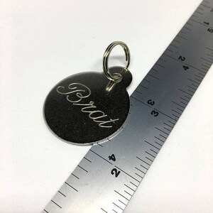 May include: Silver round keychain with a ring and the word "Brat" engraved on it.