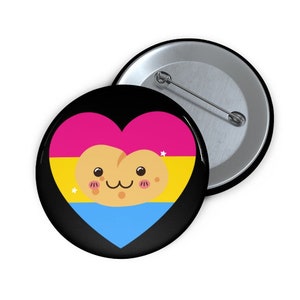May include: A black button with a heart-shaped design in the colors of the pansexual pride flag: pink, yellow, and blue. A cute, smiling cartoon character is in the center of the heart. A second button is visible in the background.