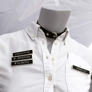 May include: A white button-down shirt with a black leather choker with a silver ring. The shirt has three black name tags with white text that read "L'AMOUR-PROPRE", "@L_AMOURPROPRE", and "@TALIMATRIX". The shirt also has a black name tag with white text that reads "MANNEQUIN".