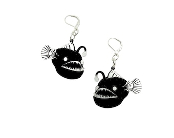 May include: A pair of black anglerfish earrings with silver-colored hooks. Each earring features a stylized anglerfish design with a wide-open mouth, sharp teeth, and a glowing lure. The earrings are set against a white background.