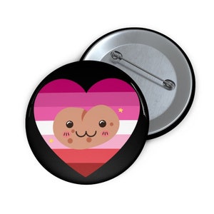 May include: A black button pin with a heart-shaped design featuring the lesbian pride flag colors: shades of pink, white, and red. A cartoon potato character with a smiling face is in the center of the heart. A second button pin is visible in the background.
