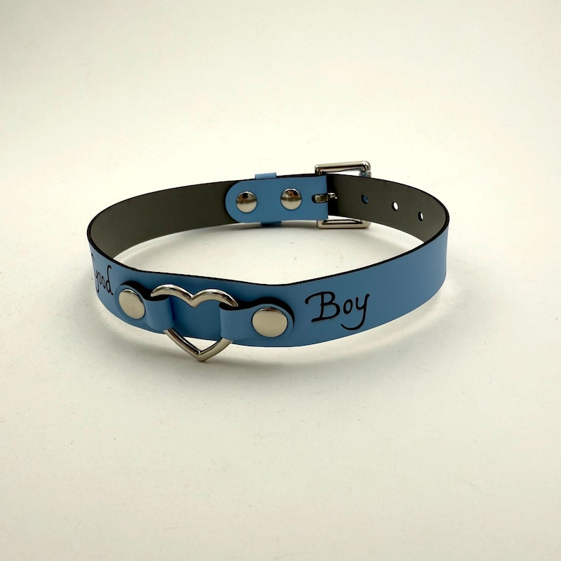 May include: A pale blue collar featuring a heart-shaped pendant and the word "Boy" in black script. It has a silver buckle and rivets. The interior is grey.