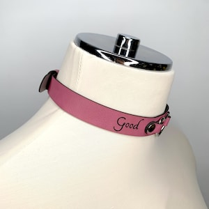May include: A pink leather choker with silver hardware and the word "Good" engraved on the front.