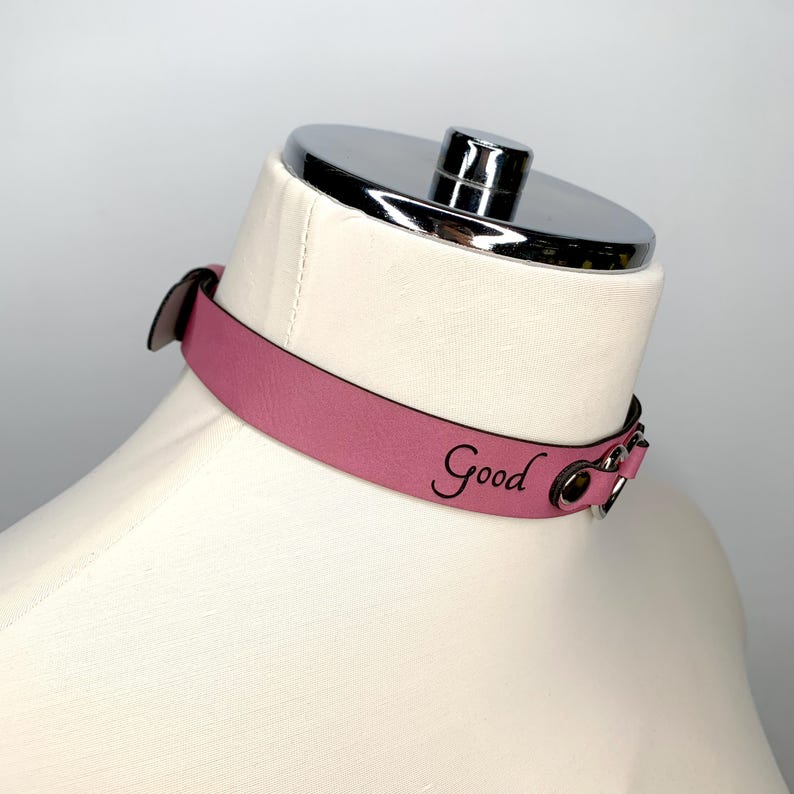 May include: A pink leather choker with silver hardware and the word "Good" engraved on the front.