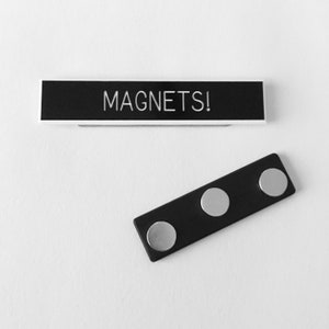 May include: A black name tag with the text "MAGNETS!" printed in white. The name tag is attached to a black magnetic strip with three silver magnets.