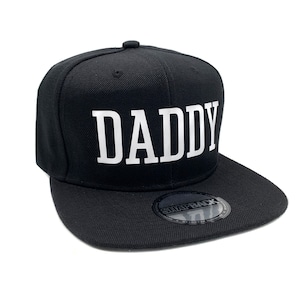 May include: Black and white baseball cap with the word "DADDY" in white lettering on the front. The cap has a black snap closure.