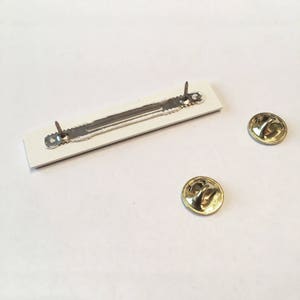 May include: White rectangular pin back with a silver metal bar and two silver pins. Two gold metal pin backs are also shown.