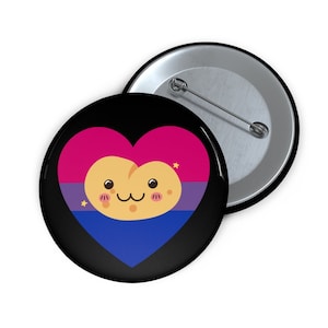 May include: A black button with a heart-shaped design in the colors of the bisexual pride flag: pink, purple, and blue. A cute, smiling peach character is in the center of the heart. A second button is visible.