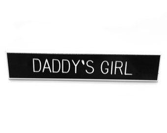 May include: Black rectangular sign with rounded corners and white text that reads "DADDY'S GIRL".