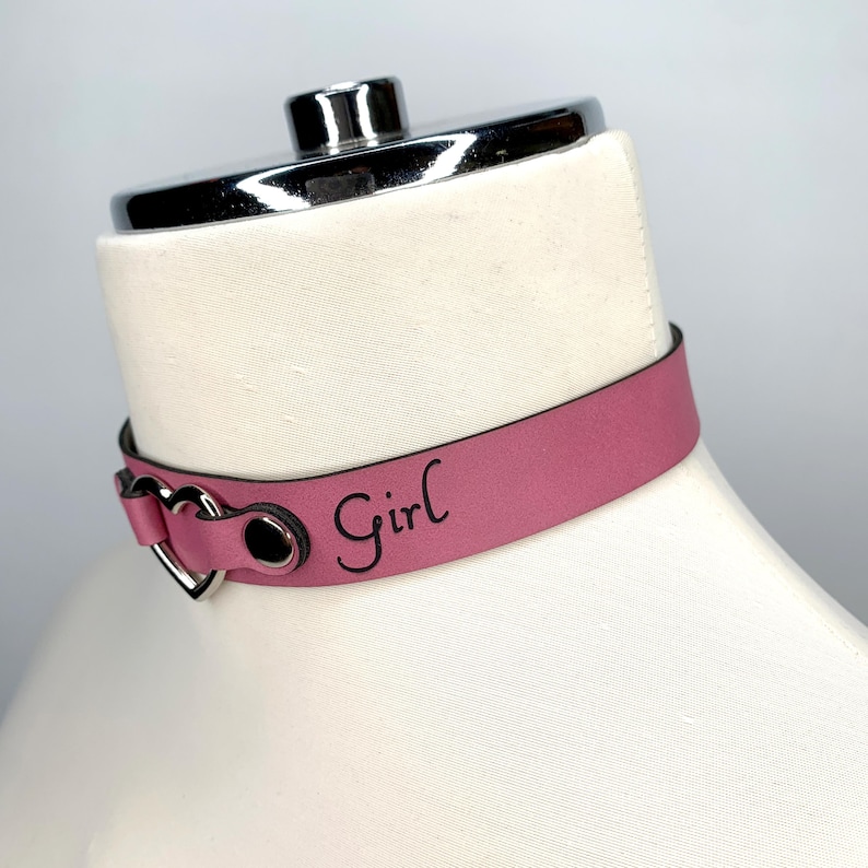 May include: A pink leather choker with a silver heart-shaped buckle and the word "Girl" engraved on the strap.