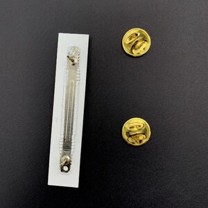 May include: The back of a white pin back with a silver clasp and two gold metal pin backs.
