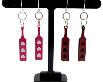 May include: A set of four dangle earrings featuring a paddle design with three hearts. Two earrings are pink with white hearts and two earrings are red with black hearts.