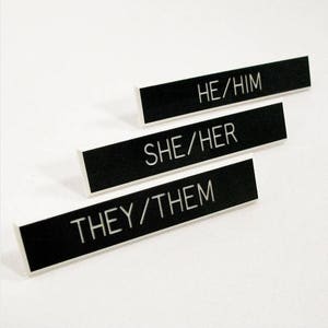 May include: Three black rectangular name tags with white text. The text on the name tags reads "HE/HIM", "SHE/HER", and "THEY/THEM".