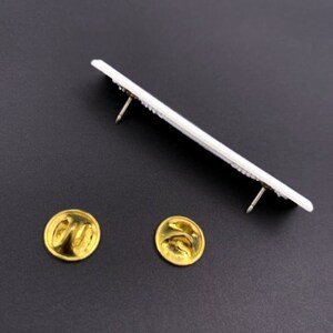 K&ouml;nnte beinhalten: Zwei goldfarbene Sicherheitsnadeln, eine links und eine rechts, sind auf einer schwarzen Oberfl&auml;che zu sehen. Eine wei&szlig;e rechteckige Nadel mit zwei goldfarbenen Sicherheitsnadeln ist &uuml;ber den beiden Sicherheitsnadeln zu sehen.