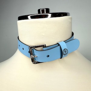 May include: A pale blue choker collar with a silver-coloured buckle and a matching strap. The collar is displayed on a white mannequin. The strap has a circular logo. The collar is made of a smooth material.