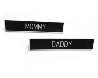 May include: Two black rectangular name tags with white lettering. The top tag reads "MOMMY" and the bottom tag reads "DADDY".