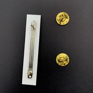 May include: A white rectangular pin back with a silver metal pin attached. Two gold metal pin backs are also shown.
