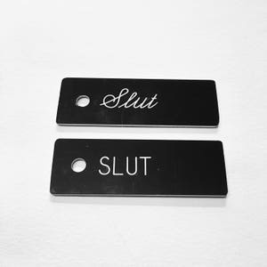May include: Two black metal tags with the word "SLUT" written on them in white. The top tag has the word written in cursive.