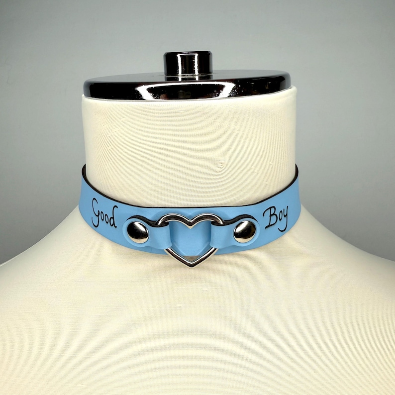 May include: A pale blue choker collar with the words "Good Boy" in black script. The collar has a silver heart-shaped centre and silver studs. The collar is displayed on a mannequin.