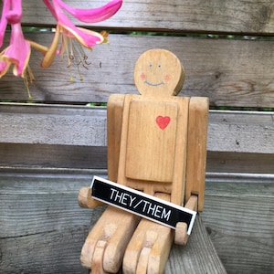 May include: A wooden toy person with a painted heart and a sign that says "They/Them". The toy is sitting on a wooden surface.