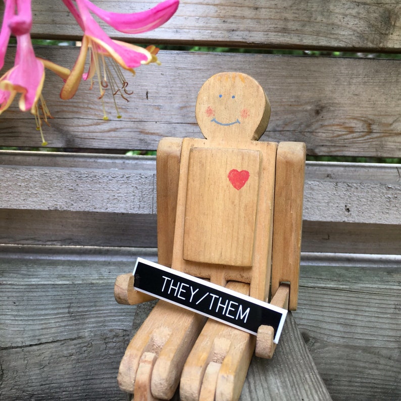 May include: A wooden toy person with a painted heart and a sign that says "They/Them". The toy is sitting on a wooden surface.