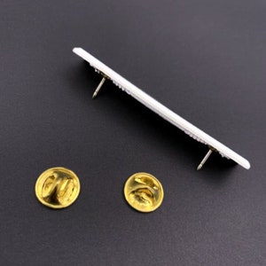 May include: Two gold-colored pin backs, one on the left and one on the right, are shown on a black surface. A white rectangular pin with two gold-colored pin backs is shown above the two pin backs.
